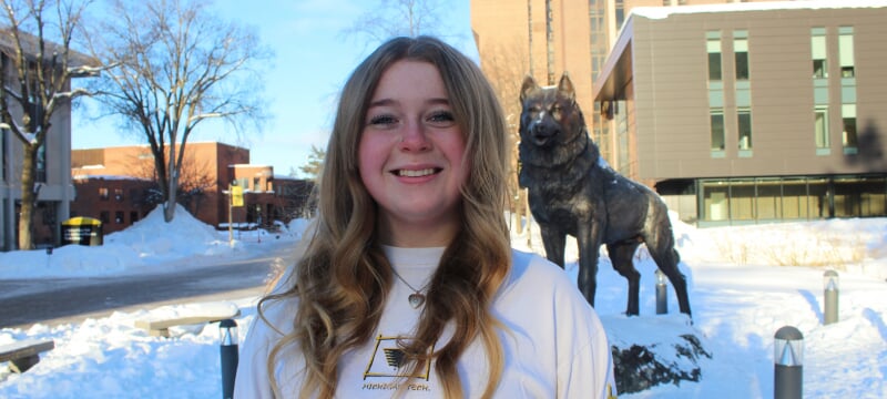 Kallee McCone in a Michigan Tech sweatshirt in front of the Husky Statue during a sunny winter day.