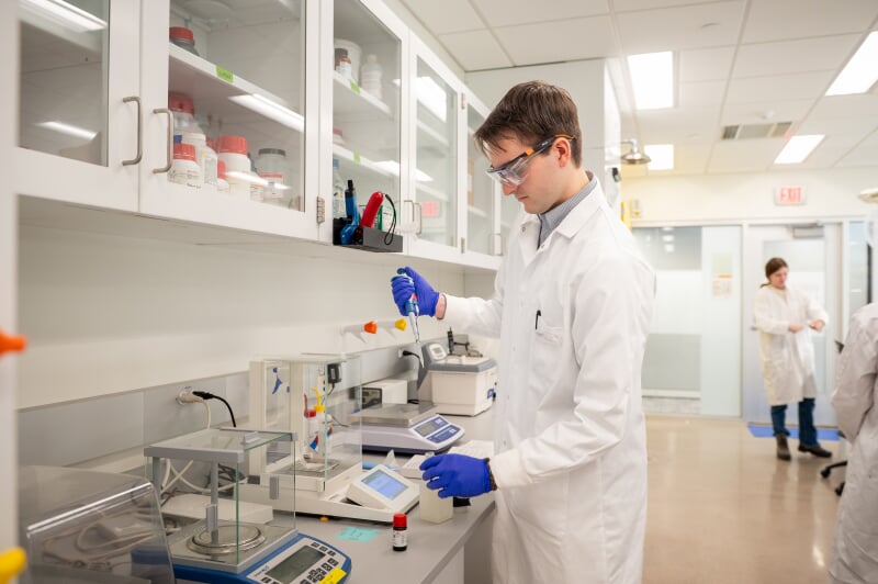 A research assistant in a white lab coat and blue gloves using a pipette to transfer liquid in a modern laboratory setting.