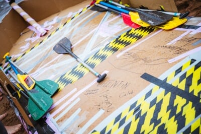 The interior of a cardboard boat with paddles marked 'Homecoming,' and the signatures of student racers, including Valentin, Caleb, Nathan, and Vincent.