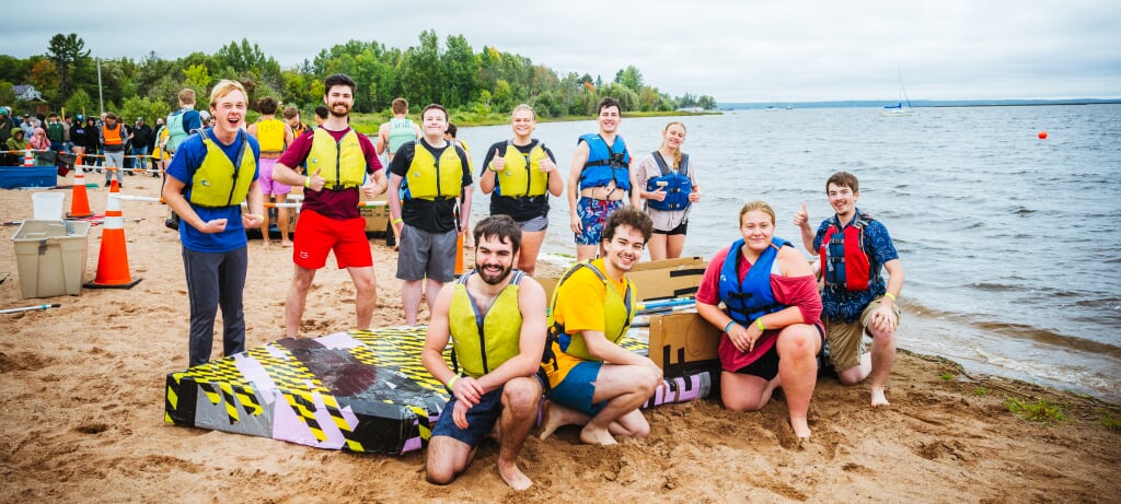 Michigan Tech's East Hall crew is ready to race. Ten Michigan Tech students pose on a beach with their cardboard boat, as other teams stage their boats in the background and race organizers survey the scene. There are sailboats moored in water in the background, along with a large orange buoy.