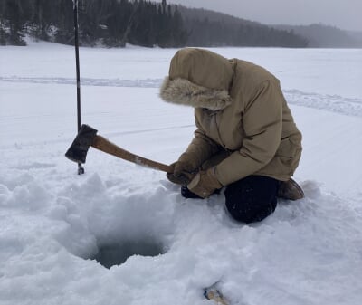Person covered by a hooded parka holding an axe and kneeling by a hole chopped in the ice.