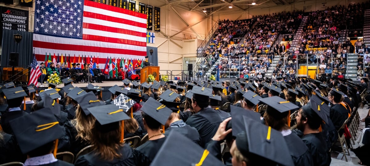 Michigan Tech’s 2026 Spring Commencement will honor more than 1,300 graduates. Looking toward the giant American flag behind the stage with many graduate in caps and gowns seated in front and people in the stands at commencement.