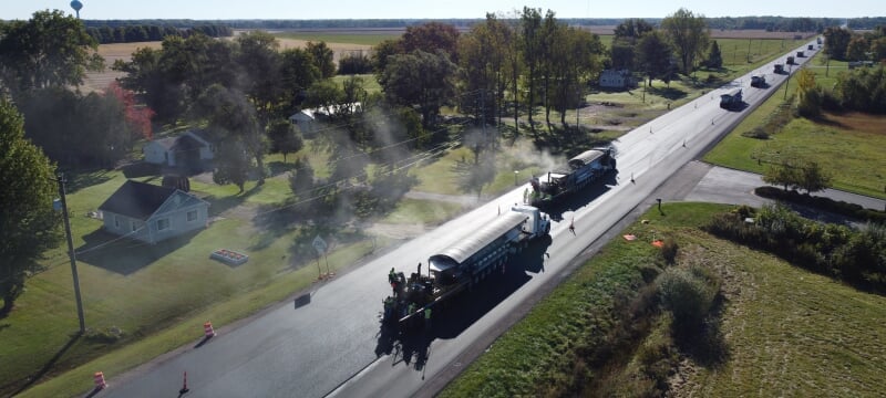 Ten trucks lay new pavement on a road through flat farmland in an aerial shot.