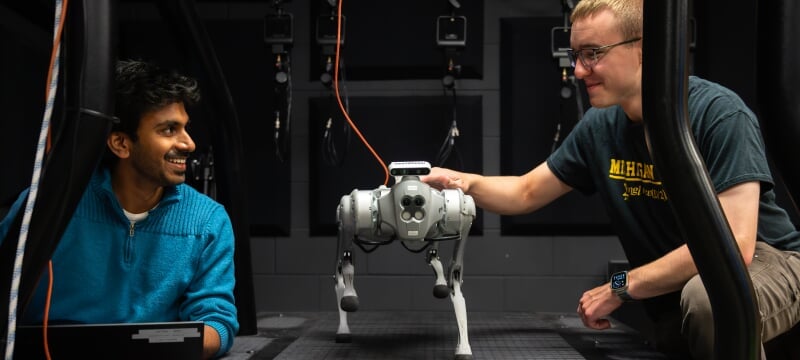 Two researchers test a quadruped robot walking on a treadmill in a lab setting.