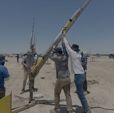Two Michigan Tech students in hats prepare a large model rocket for launch in a dry, open field under a clear blue sky.