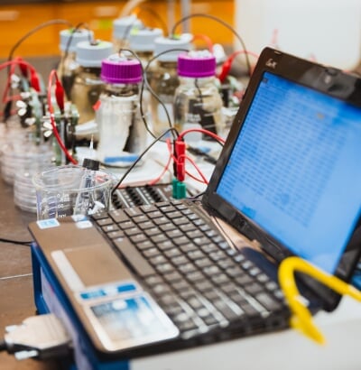 A small laptop displays a spreadsheet with data and measurements from a series of small glass bottles being monitored with scales and electrodes.