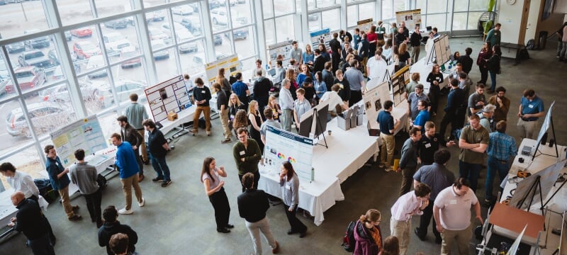 Large group of people attending an academic poster presentation in a spacious, sunlit hall with floor-to-ceiling windows.