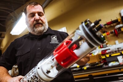 Jamey Anderson stands in a lab, wearing a shirt with the Michigan Tech Great Lakes Research Center logo over the left breast pocket, holding a long cylindrical component containing pumps for the MFC.