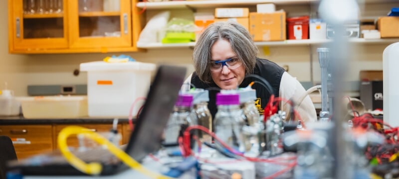 Amy Marcarelli stands in a lab wearing safety goggles, leaning over to get a closer look at glass bottles being monitored for data by a laptop and electrodes.