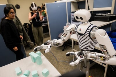 A student wearing a VR headset directs Reachy the Robot to grasp and pile blocks on a table while another student watches.