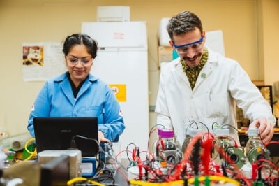 Two student researchers stand side by side in a lab, wearing lab safety goggles and lab coats. They are smiling and looking down at a table full of bottles filled with liquid, monitored by sensors feeding data into a laptop.
