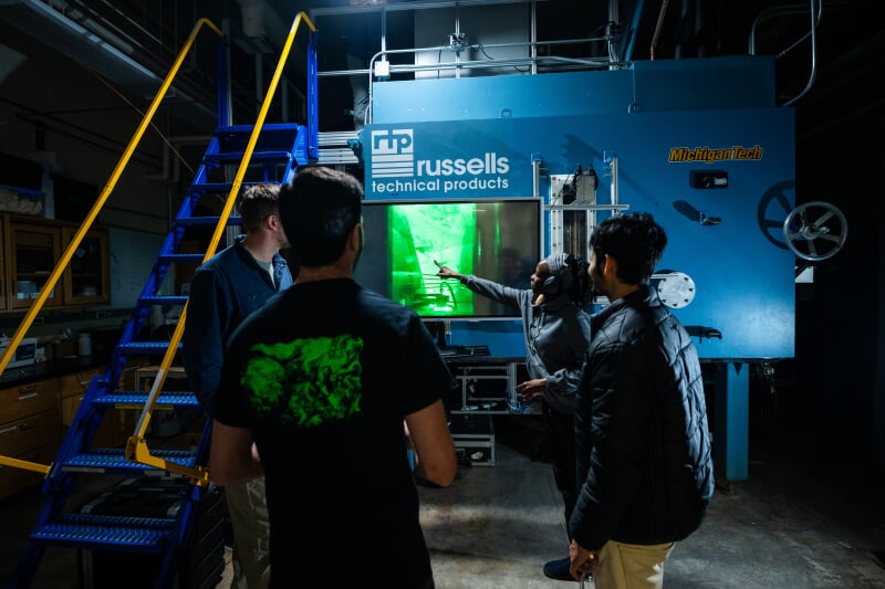 Four people stand in front of Tech’s Pi Cloud Chamber, a large blue box-like piece of equipment with a digital monitor displaying green lights that represent cloud data.