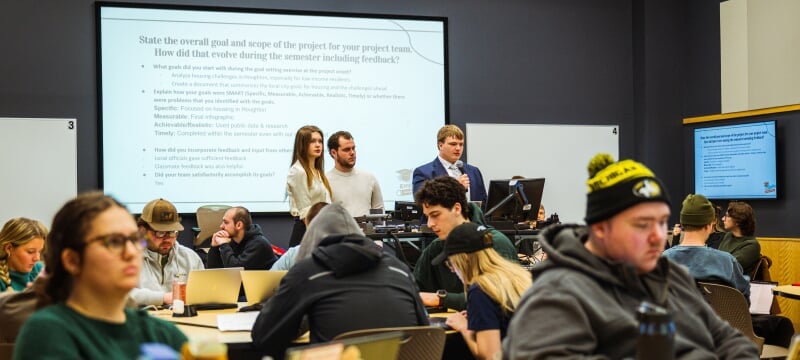 Three students share their SMART goals on a projection screen behind them as other students in the class look on.