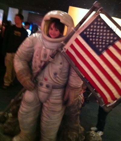 A grainy photo of a young Jennifer Celeste posing in an astronaut suit holding an American flag at the Museum of Science and Industry in Chicago, Illinois.