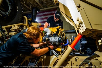 Two men work on the internal components of a large tan tactical vehicle.
