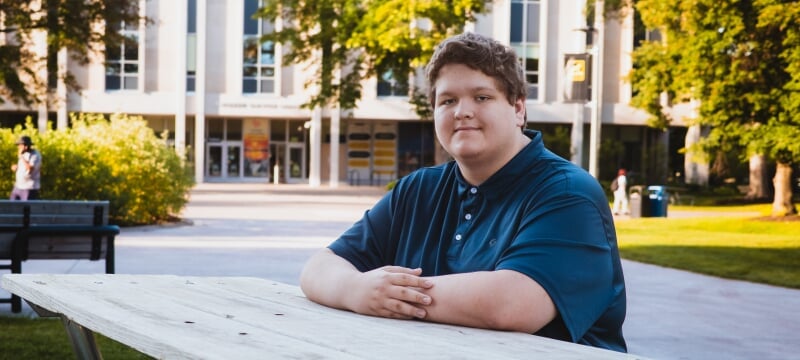 William Ingels sits at a picnic table wearing a blue polo in front of the Van Pelt and Opie Library.