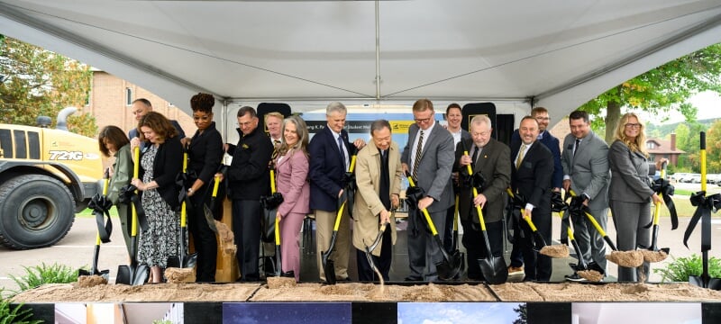 Campus leaders on a platform stage stand with black and gold shovels turning dirt during a groundbreaking ceremony. 