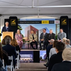 President Koubek speaking at the podium while the others on stage watch him during the groundbreaking ceremony.