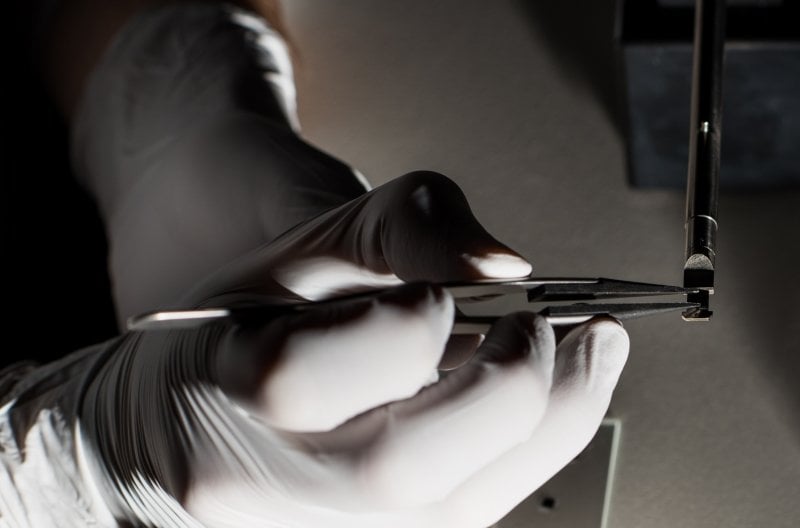 Gloved hands holding fine-tipped tweezers place a rice grain-sized sample holder in place on a pencil-sized instrument