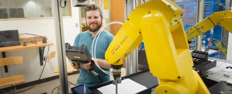 A young male student stands in a testing lab near a robotic machine indoors in a university lab