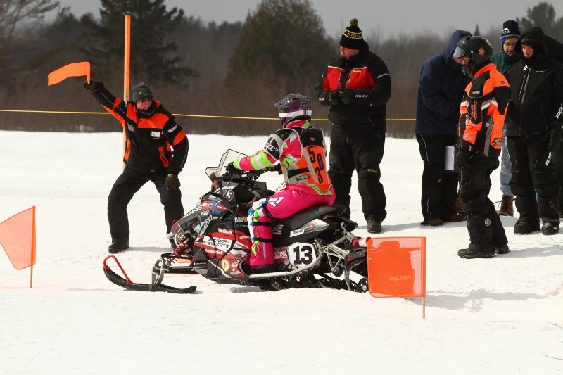 A man raises an orange flag in front of a snowmobile rider waiting to start a race.