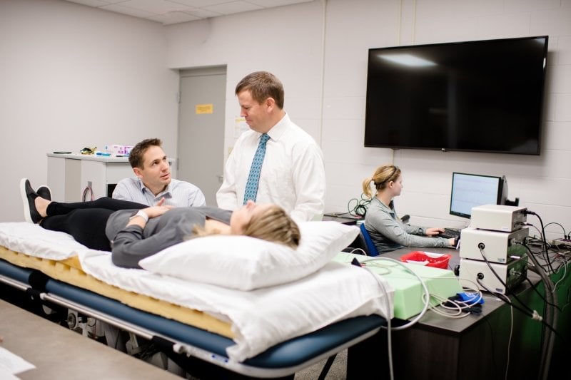 researcher stand next to a young woman laying on a bed in a lab