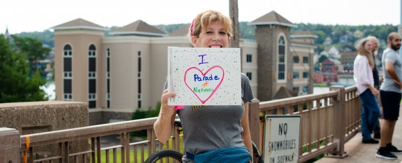 woman on a bridge outside with the sky behind her holding a sign that says I heart Parade of nations