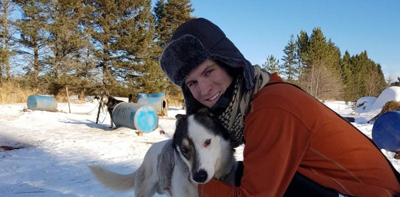 A young man is a fur hat with his sled dog outside with blue barrels on a snowy day in the dog kennel area