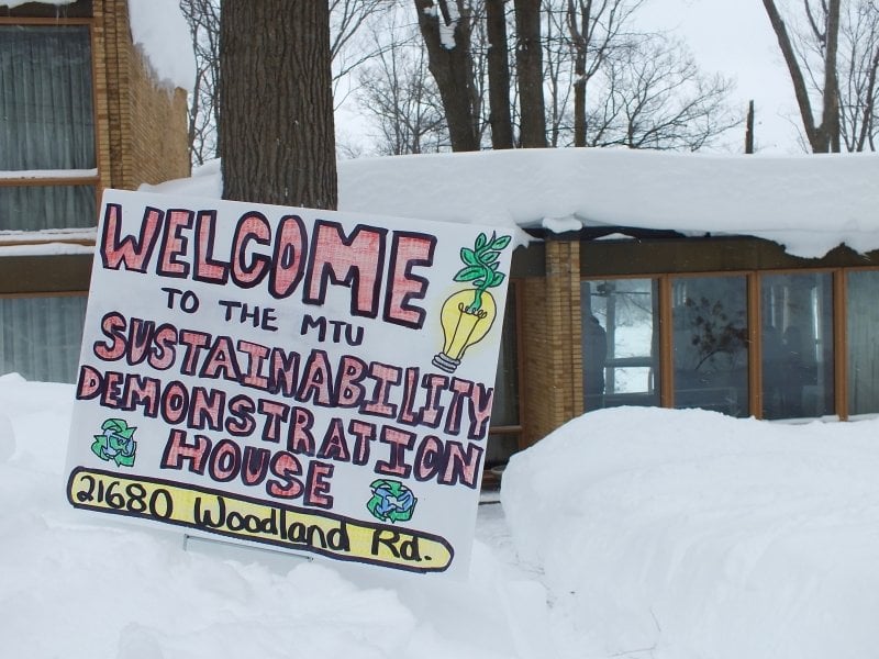 Tan brick house with an open house sign propped on a tree reading 21680 Woodland Road in the snow in winter.