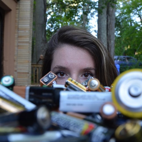 a woman's face behind a pile of collected batteries with a brick building and woods in the background