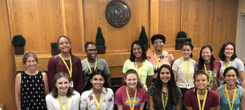 A woman in a polka dot dress with 13 students in summer clothes in front of a fireplace and wood paneling with the Michigan Technological University seal above at a summer STEM workshop