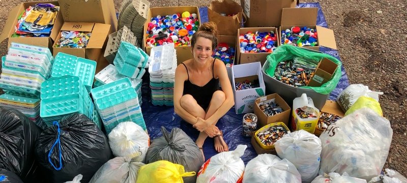 A young woman sitting in the center of recycled egg cartons, foil-lined wrappers, bottle caps, plastic bags and batteries.