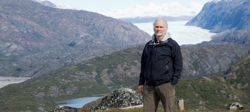 A man stands in front of mountains with green moss, blue sky, snow caps and water in the distance