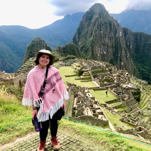 A young woman in a pink poncho, hat, and hiking boots smiles above Machu Picchu in Peru with the Andes Mountains in the background.