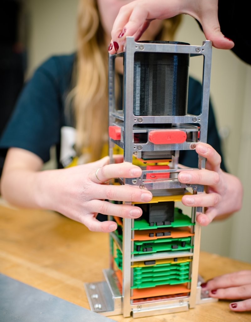 Hands hold a metal and plastic cubesat in a laboratory with a blurred background.