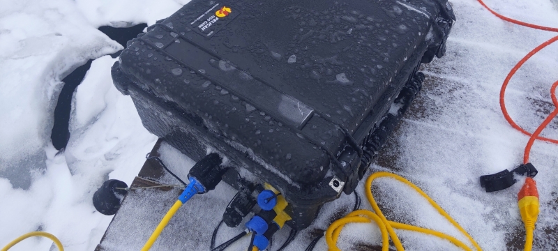 Test equipment in a black box on a snow-covered dock next to the ice in winter with yellow cords and red and black plugs