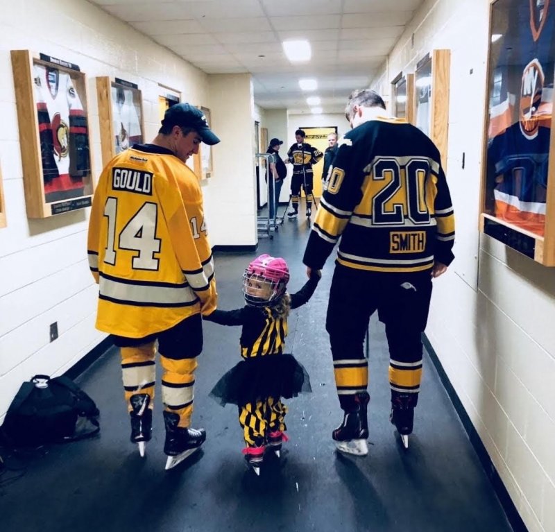 A little girl in a tutu holds the hands of two hockey players in the hall with their backs to us in Michigan Tech's John MacInnes student ice arena.