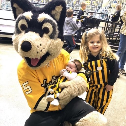 A giant Husky in a Michigan Tech jersey holds a tiny baby in striped overalls while a little gear also in striped overalls stands nearby at a Michigan Tech Hockey game in an ice arena