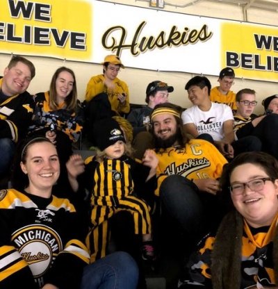 A group of students in Michigan Tech Huskies gear and a little girl also in her gear sit in a hockey arena below a Michigan Tech Believe sign.