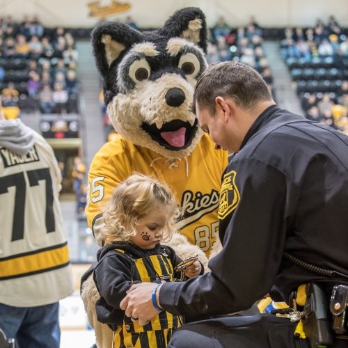A little girl holds the handcuffs of a kneeling police officer with a giant Husky mascot looking on in an ice rink at a hockey gsme in the stands.