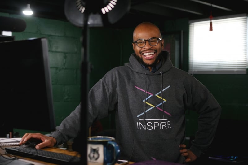 A young man wearing an Inspire sweatshirt smiles wearing glasses near a computer with a coffee cup