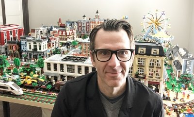 A man wearing glasses in front of a LEGO city he has constructed.