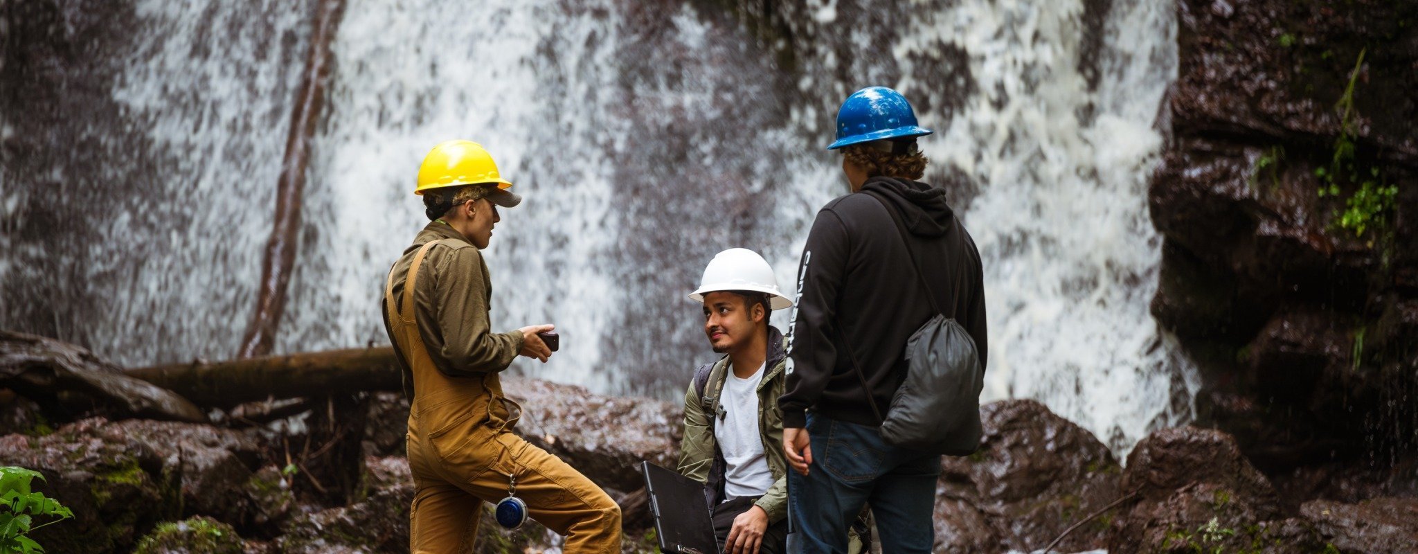 Three Michigan Tech students in the field, near a waterfall, wearing hard hats, conduct hands-on field work with measuring instruments and a laptop.