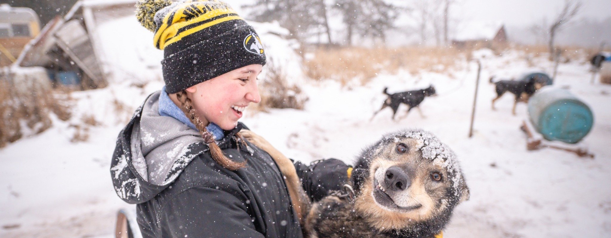 A Michigan Tech mushing club student member holds a smiling sled dog outside at the training center