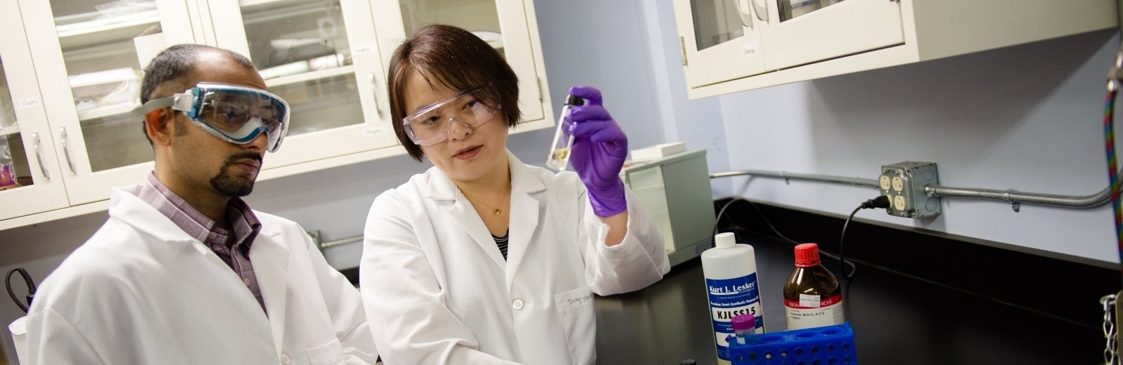 Two researchers in a lab looking at a vial of solution one researcher is holding up.