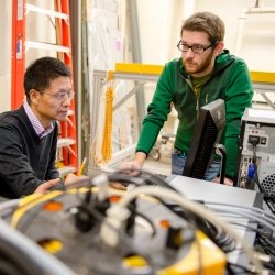 Graduate student standing over computer while another sits inside the Cloud Chamber laboratory