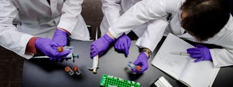 Three researchers in lab coats, gloves, and safety glasses sitting at a table with vials