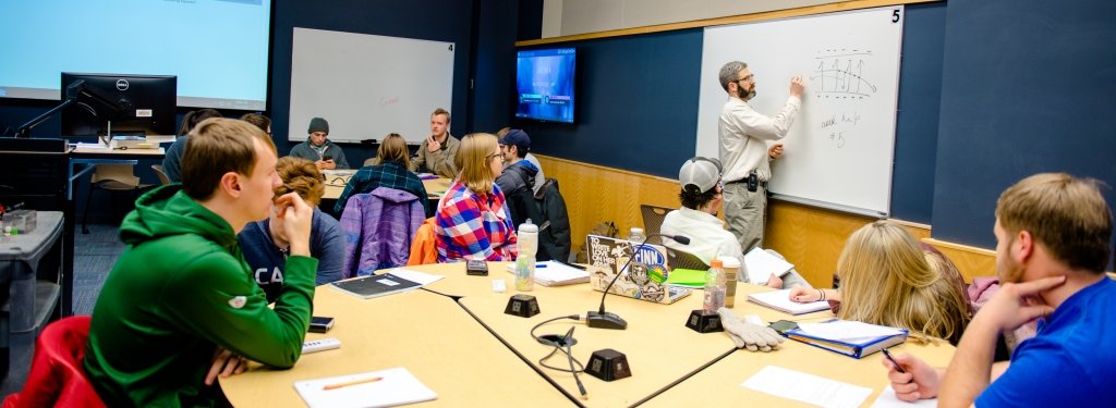 Faculty standing in front of a white board writing in front of circle tables of students looking on.