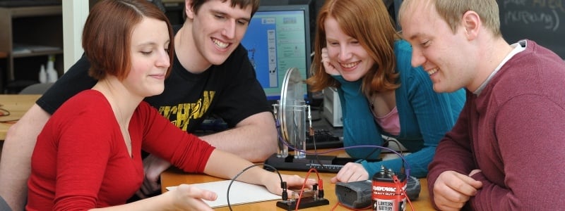Four students surrounding an experiement with a battery connected to electrodes.