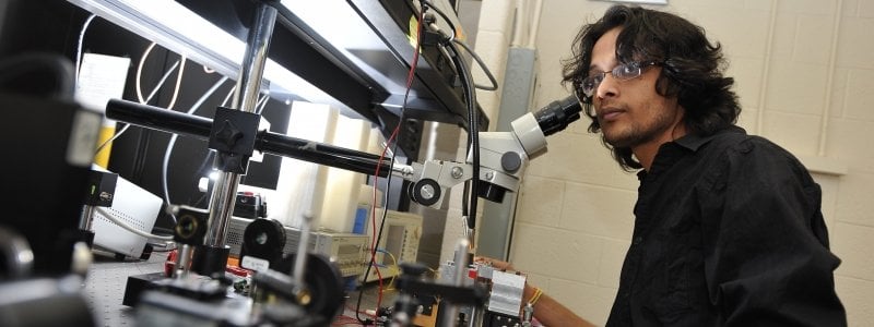 Student standing in front of a microscope on a lab table looking towards someone.